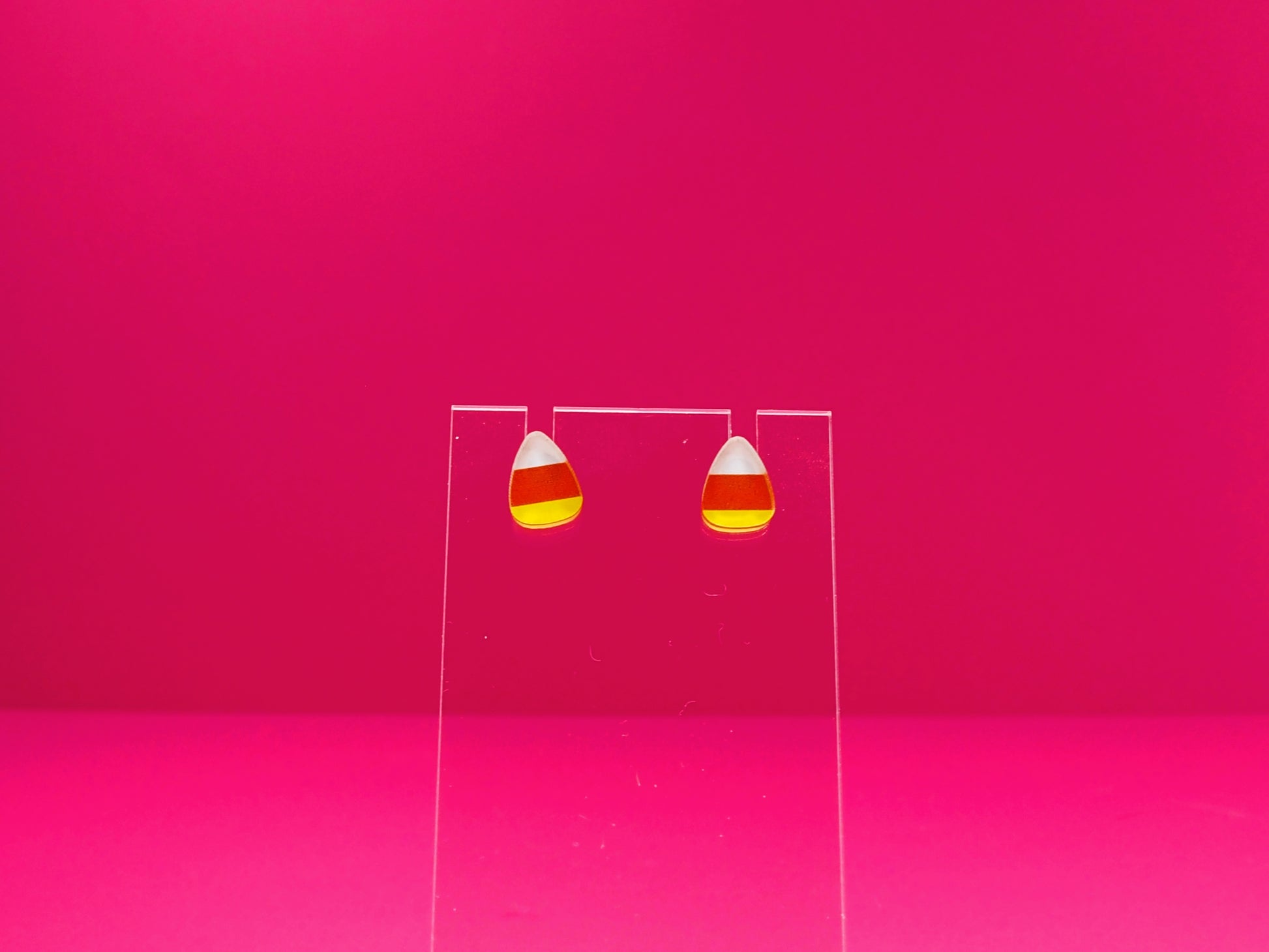 Candy corn earrings on a clear display against a pink background.