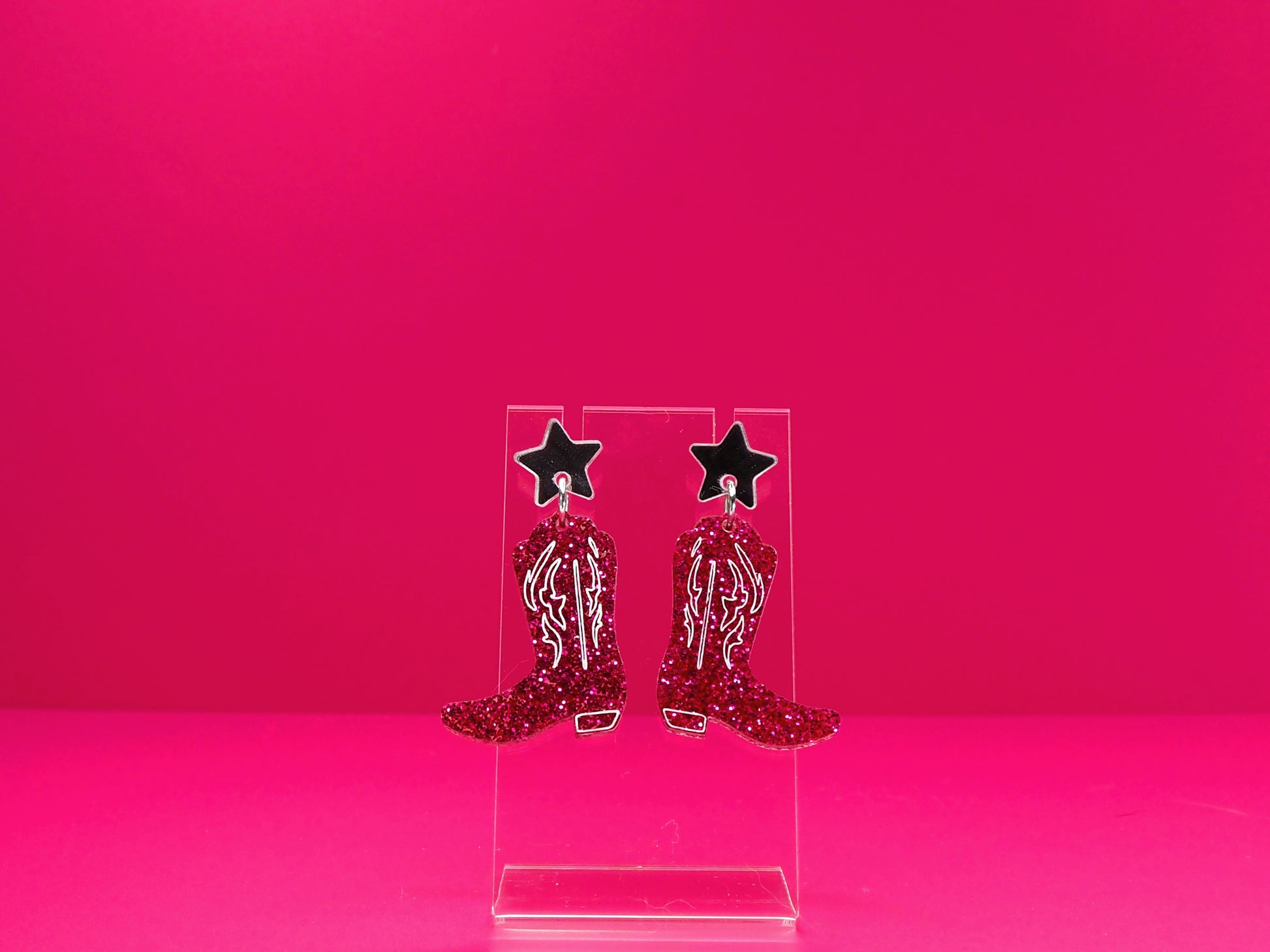 Pink cowboy boot earrings on a clear display against a pink background.