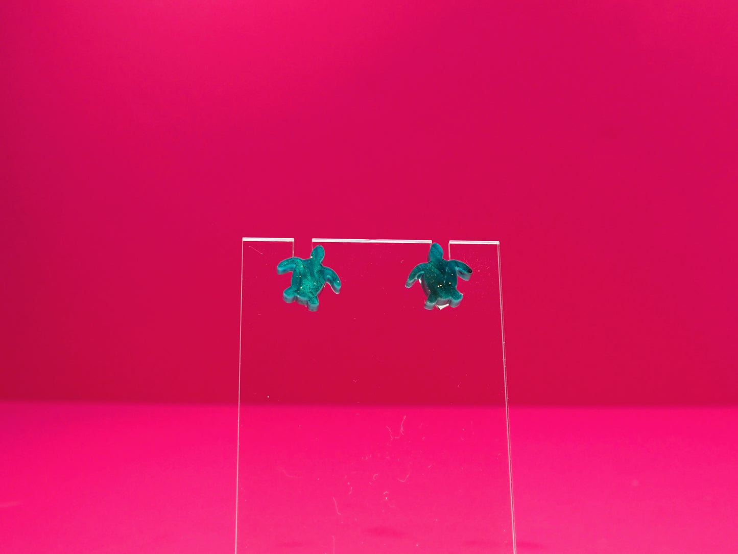 Marbled green sea turtle earrings on a clear display against a pink background.