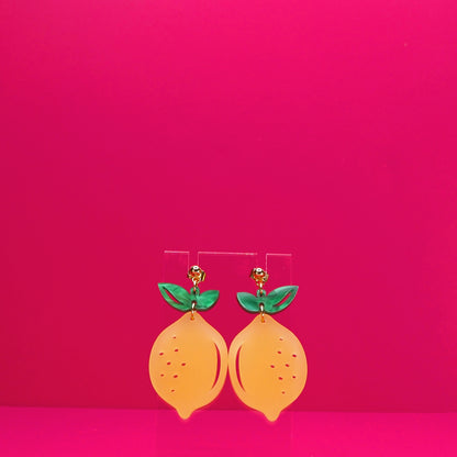 Lemon earrings on a clear display against a pink background.