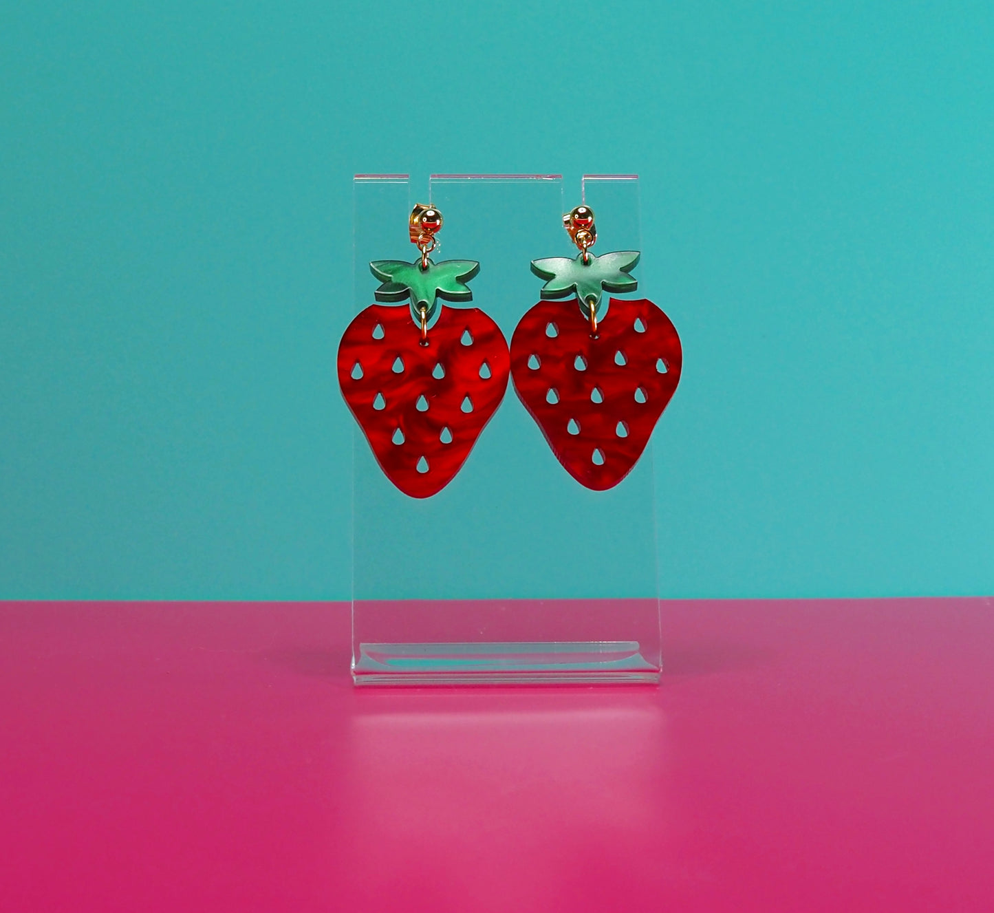 Strawberry earrings on a clear stand against a blue and pink background.