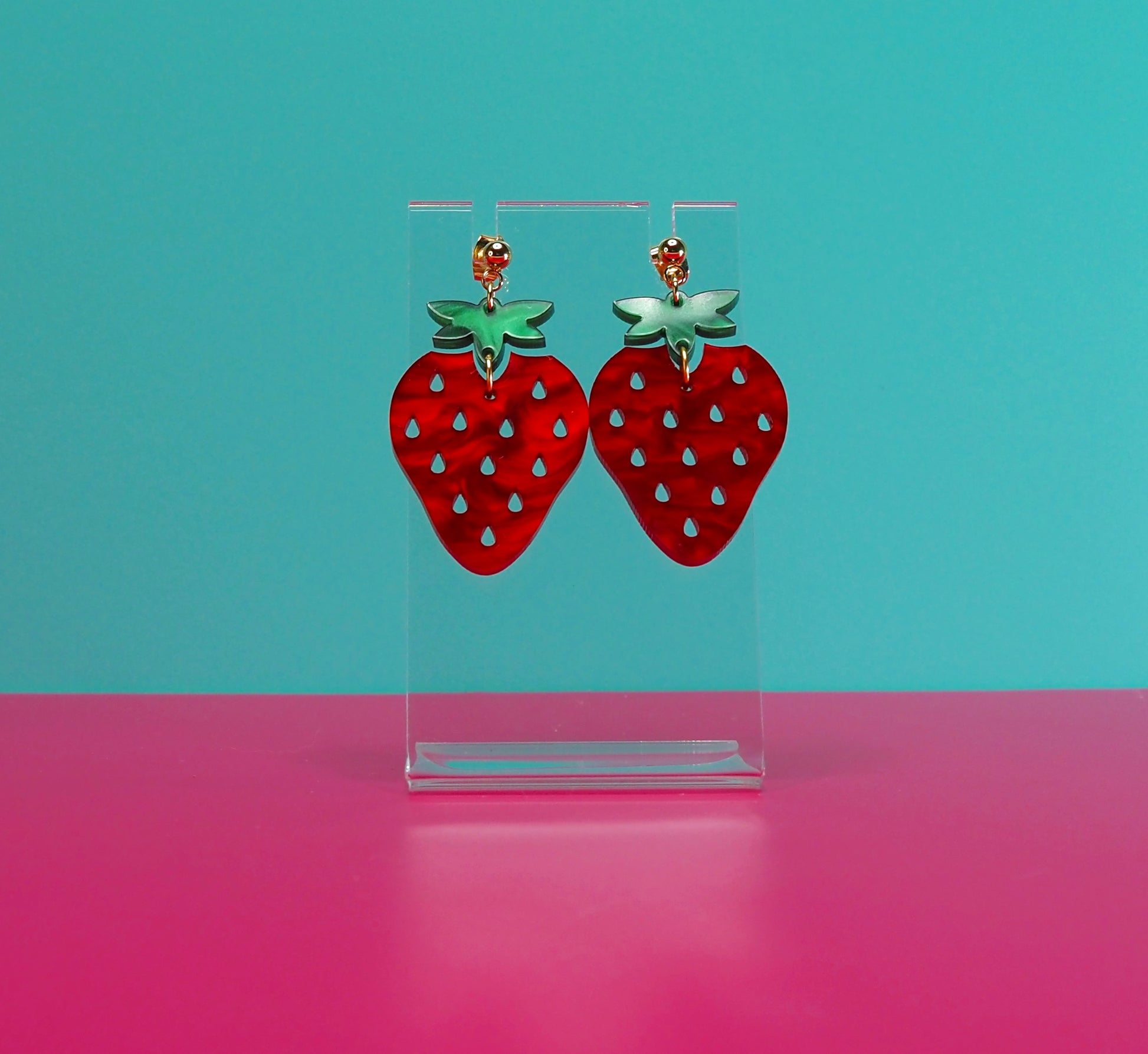 Strawberry earrings on a clear stand against a blue and pink background.