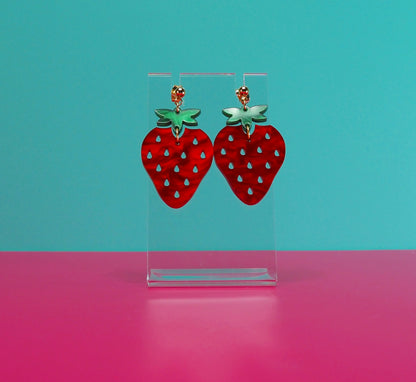 Strawberry earrings on a clear stand against a blue and pink background.