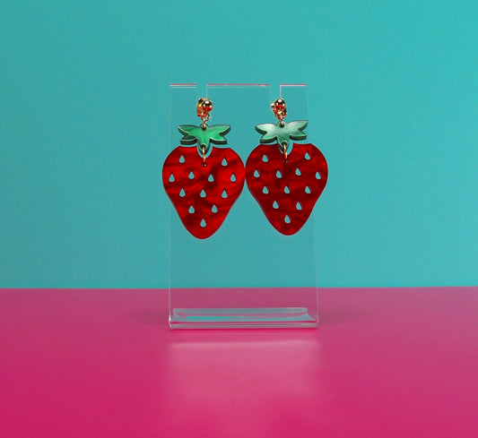 Strawberry earrings on a clear stand against a blue and pink background.