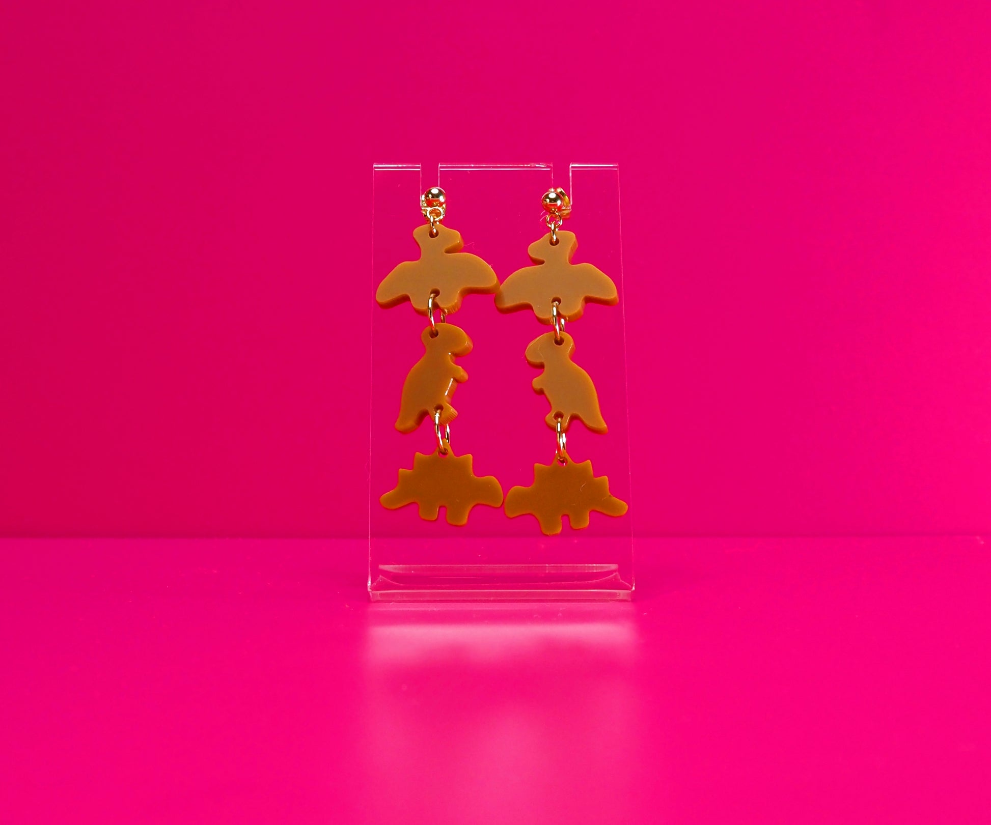 Dinosaur nugget earrings on a clear stand against a pink background.
