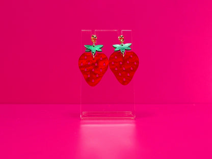 Strawberry earrings on a clear stand against a pink background.