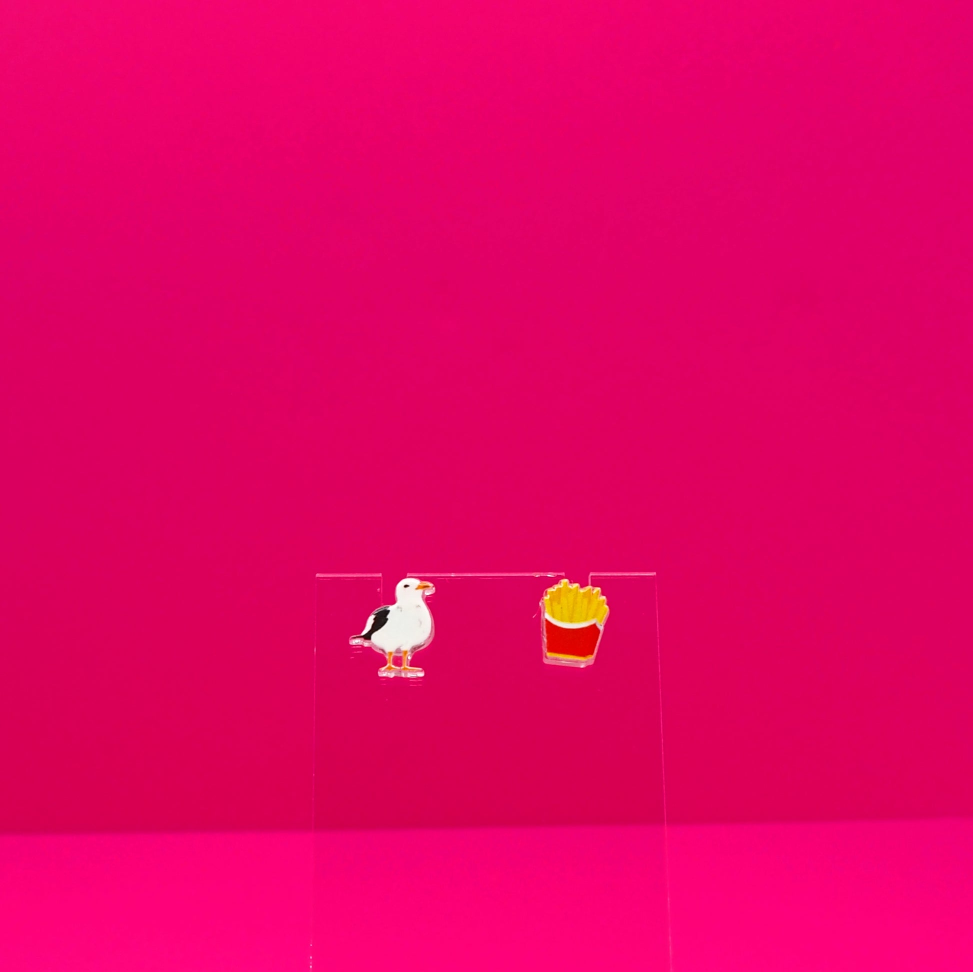 A seagull and a pack of French fries earrings on a clear stand against a pink background.