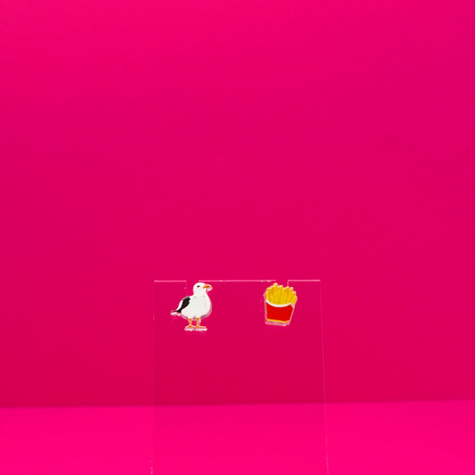 A seagull and a pack of French fries earrings on a clear stand against a pink background.