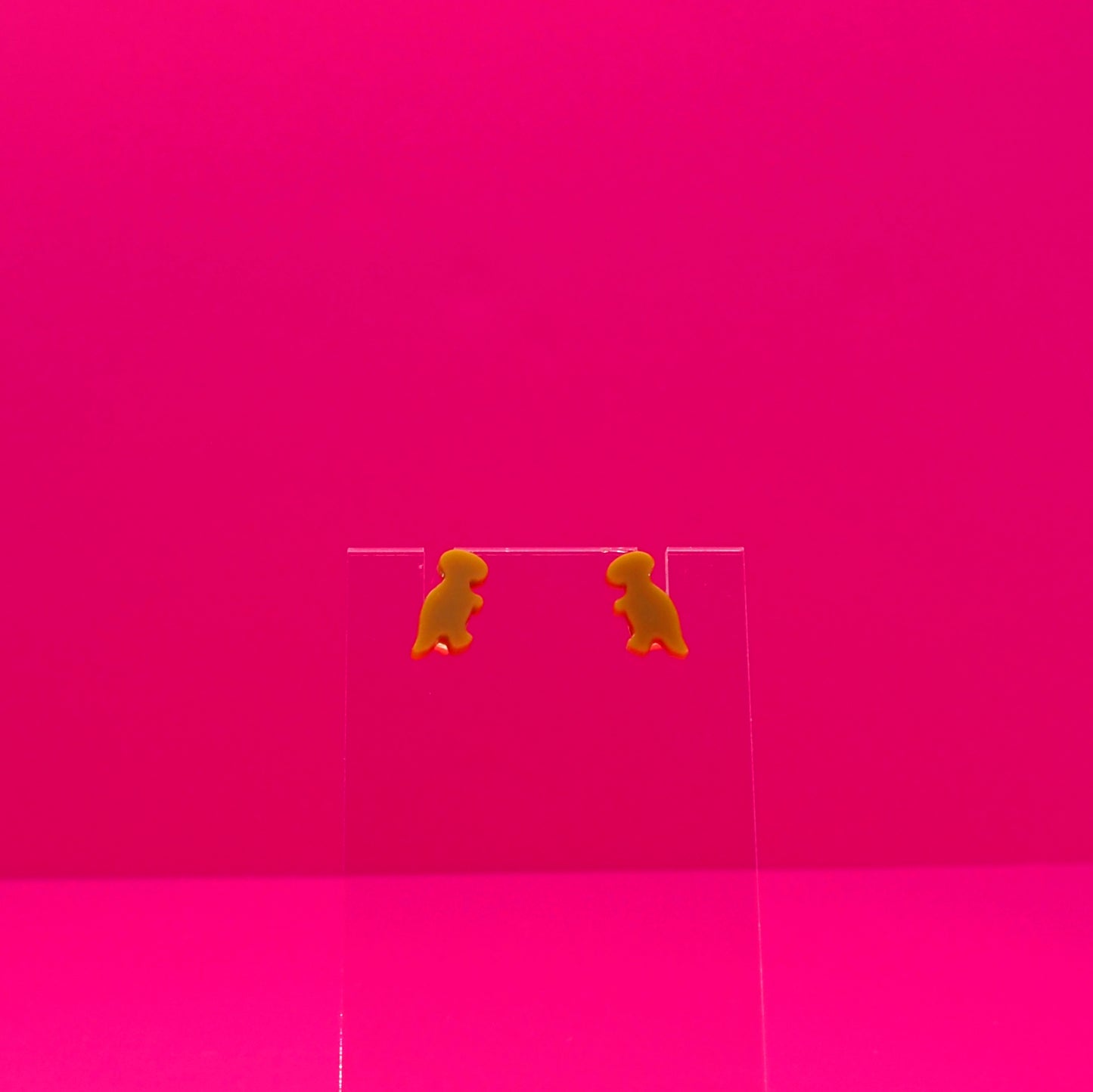 T-Rex earrings on a clear stand against a pink background.