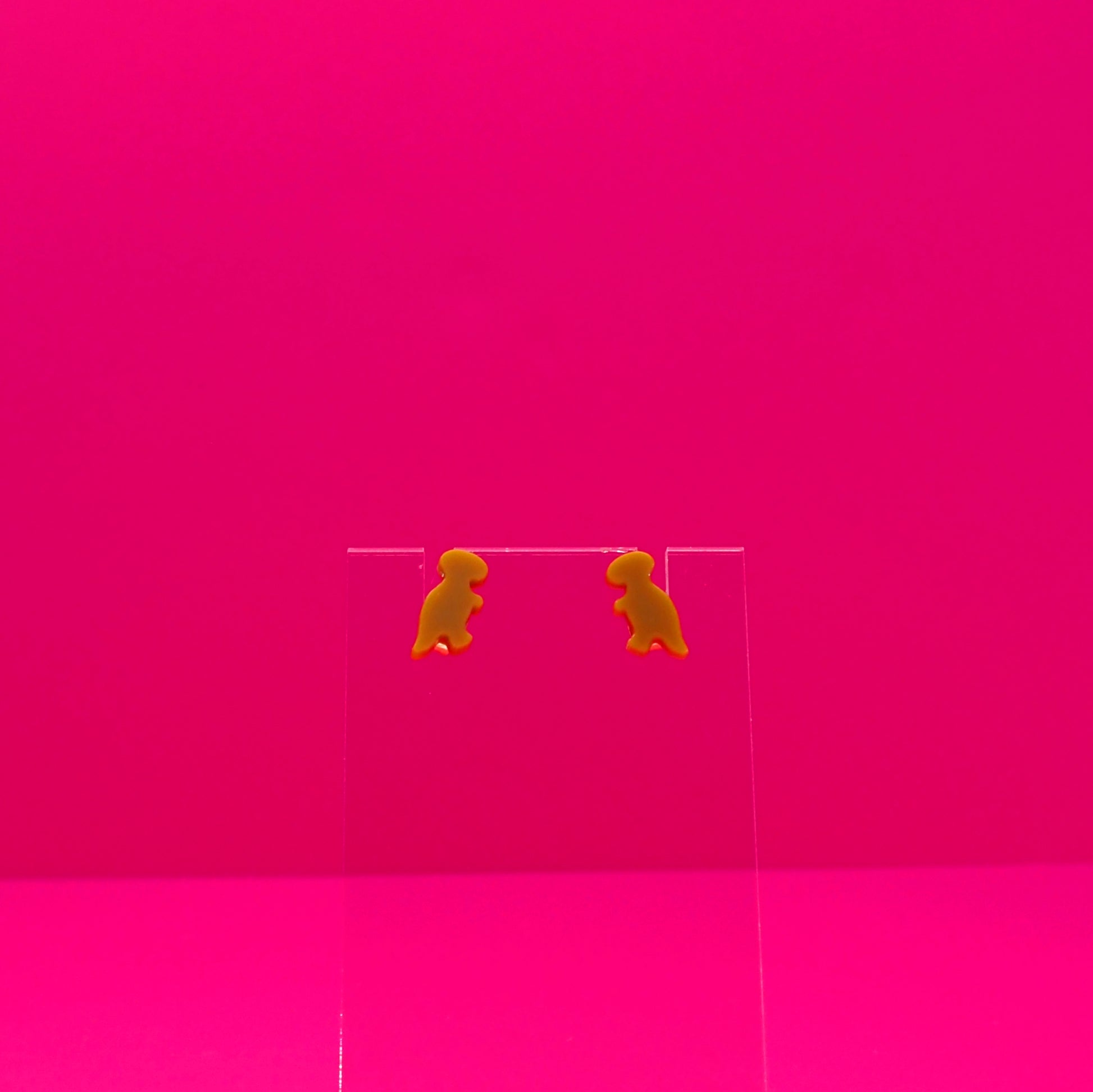 T-Rex earrings on a clear stand against a pink background.