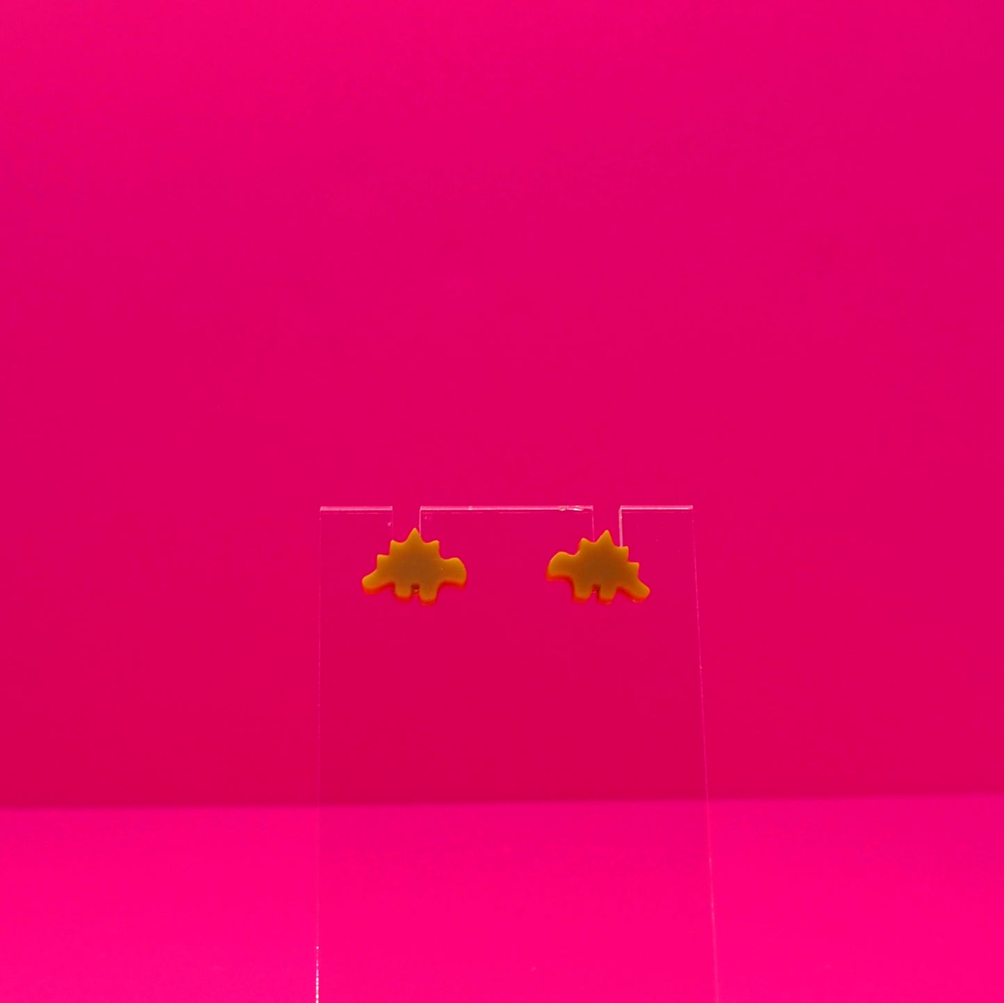 Stegosaurus earrings on a clear stand against a pink background.