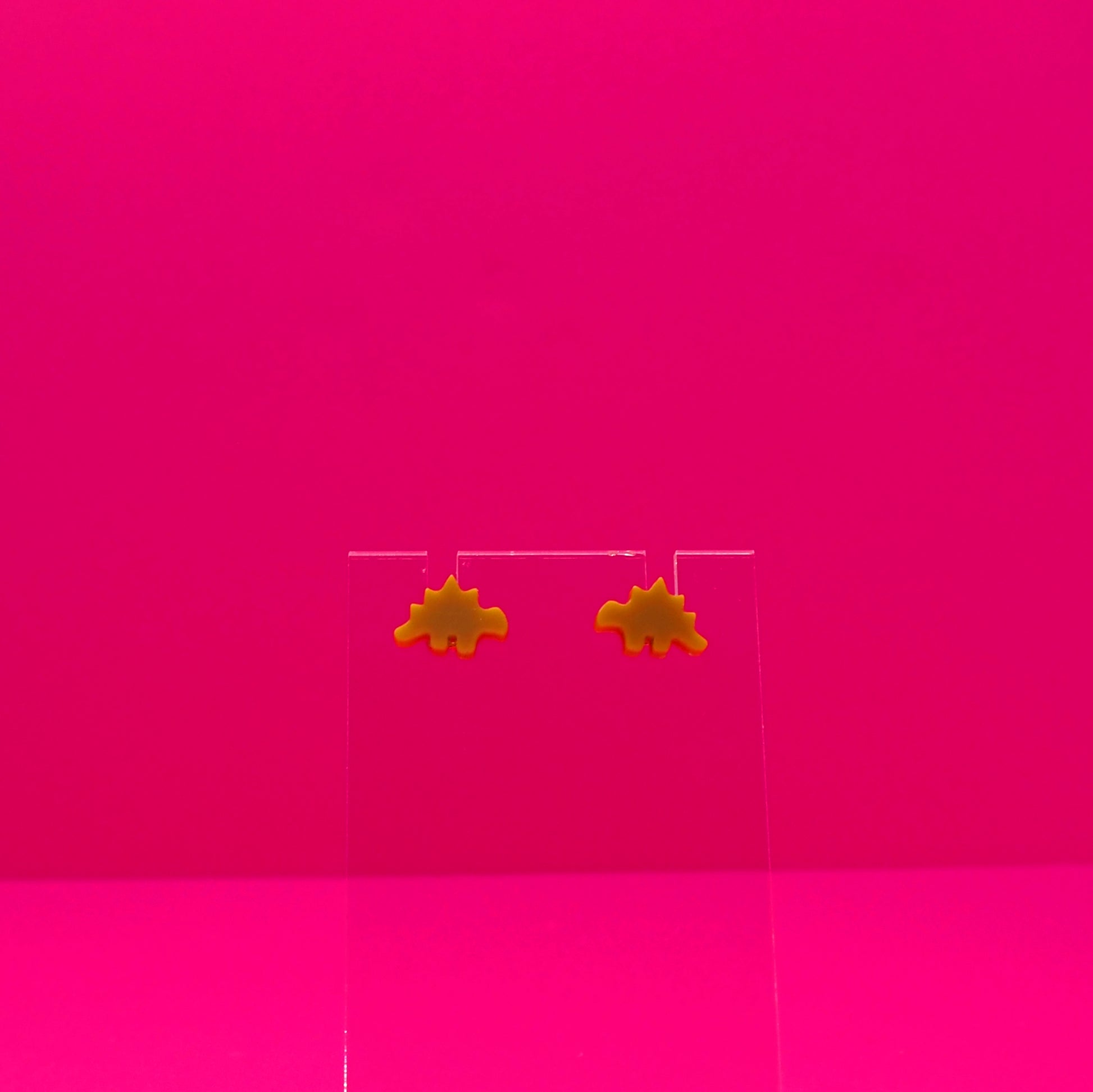 Stegosaurus earrings on a clear stand against a pink background.