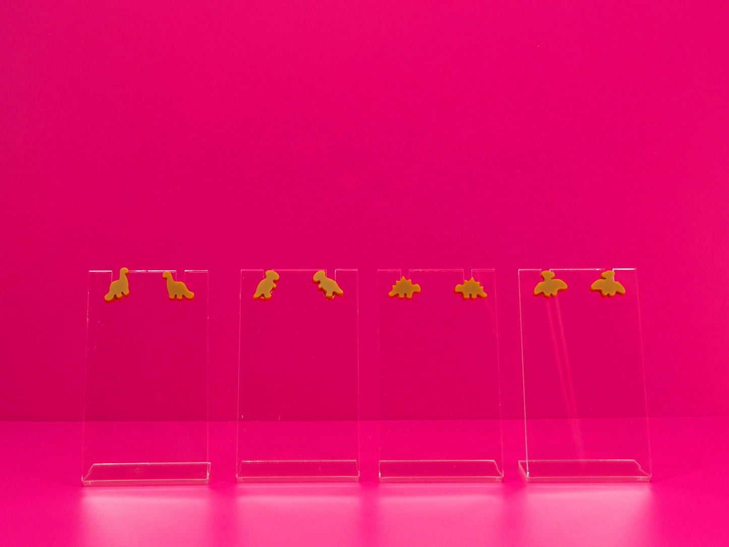 Set of brown dinosaur earrings on clear stands against a pink background