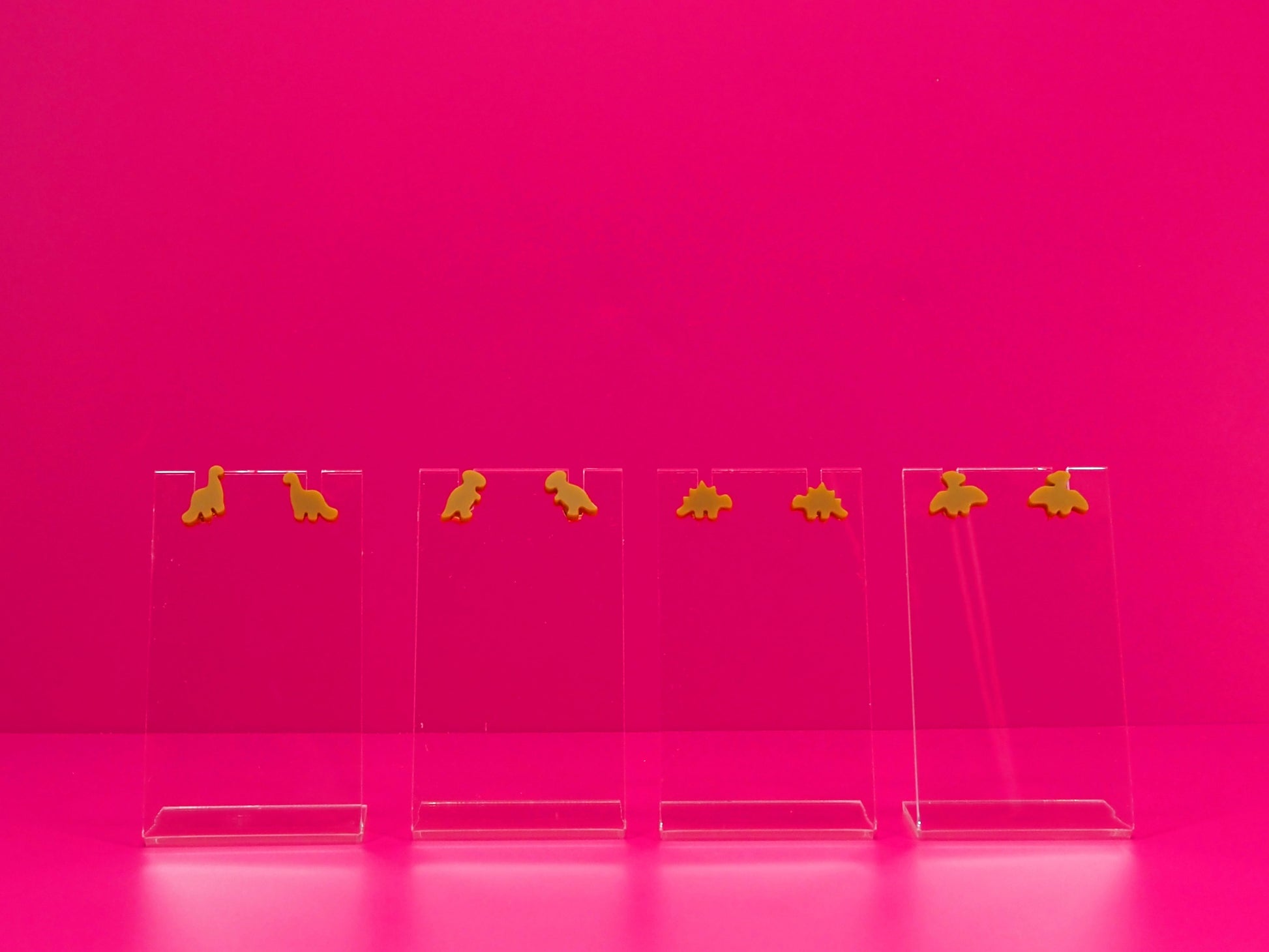 Set of brown dinosaur earrings on clear stands against a pink background