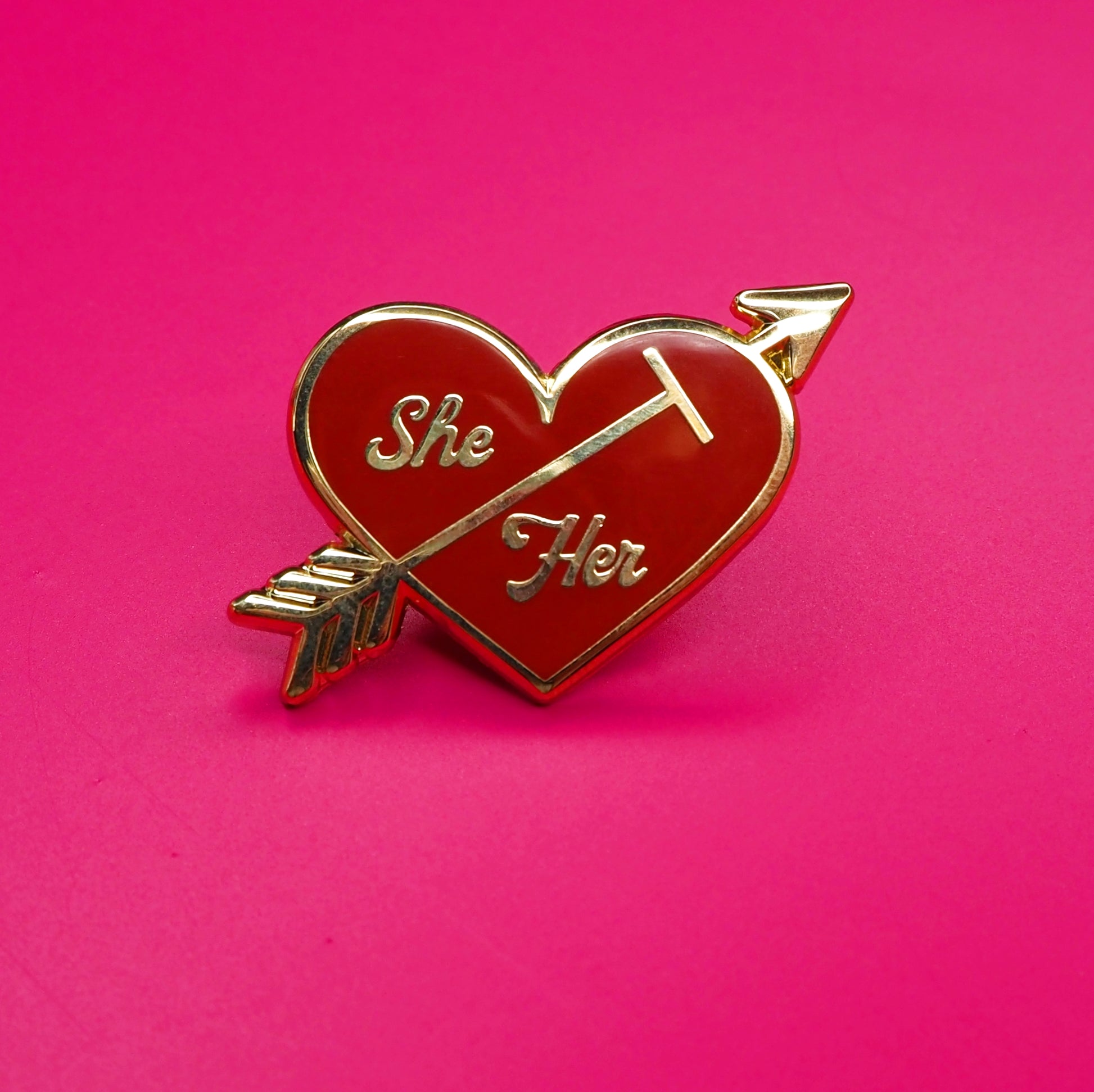 Heart-shaped pin with 'She' and 'Her' text on a pink background.