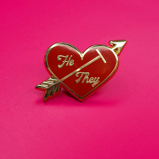 Heart-shaped pin with 'He' and 'They' text on a pink background.