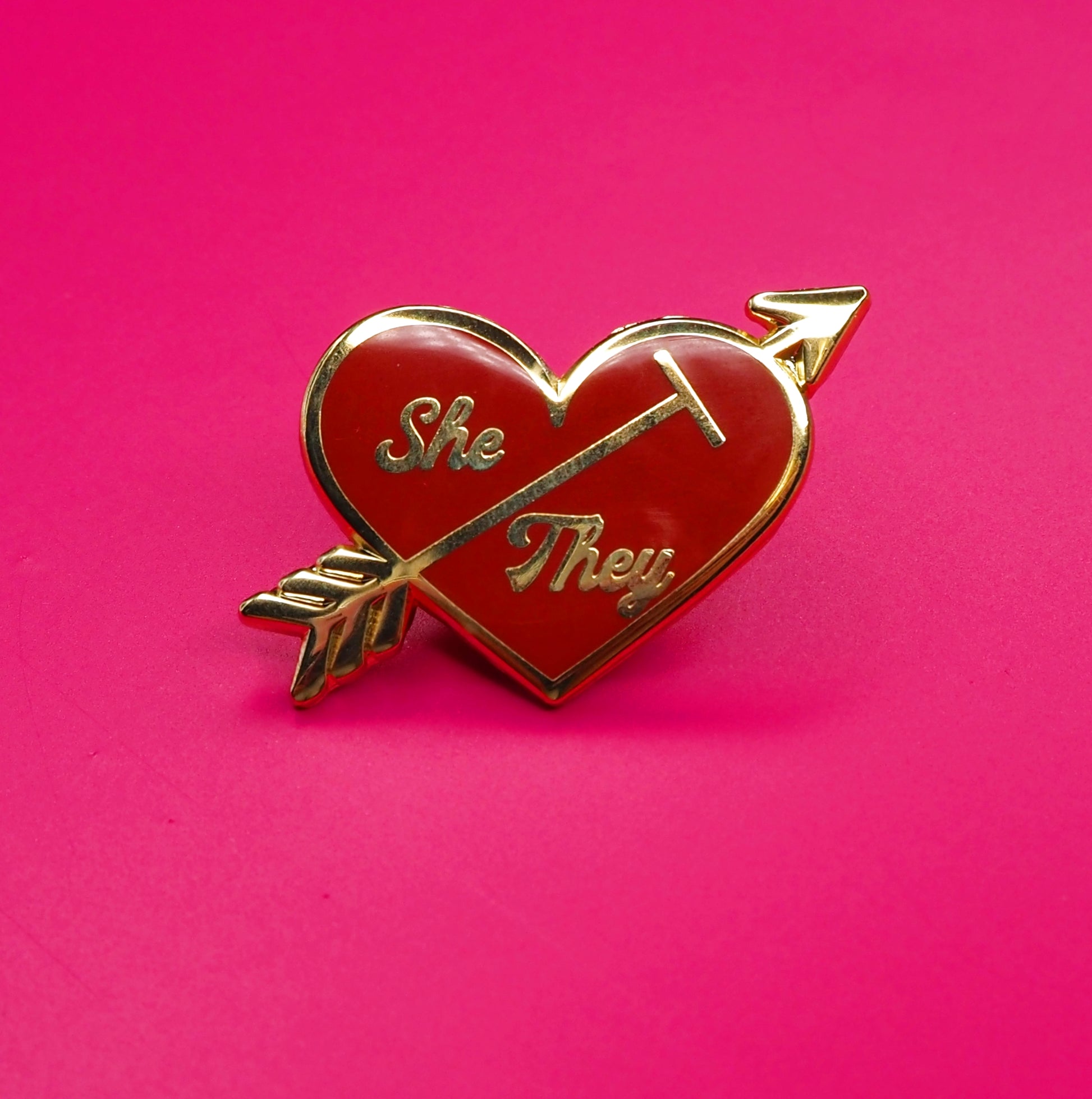 Heart-shaped pin with 'She' and 'They' text on a pink background.