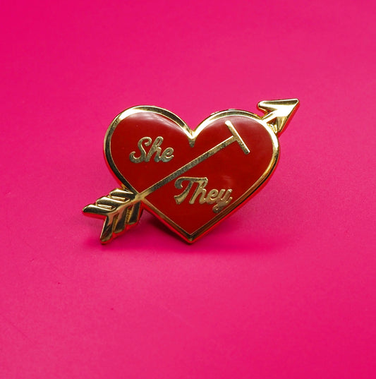 Heart-shaped pin with 'She' and 'They' text on a pink background.