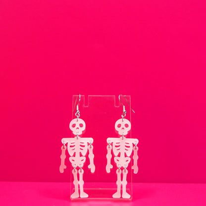 White dangling skeleton earrings on a white display against a pink background.