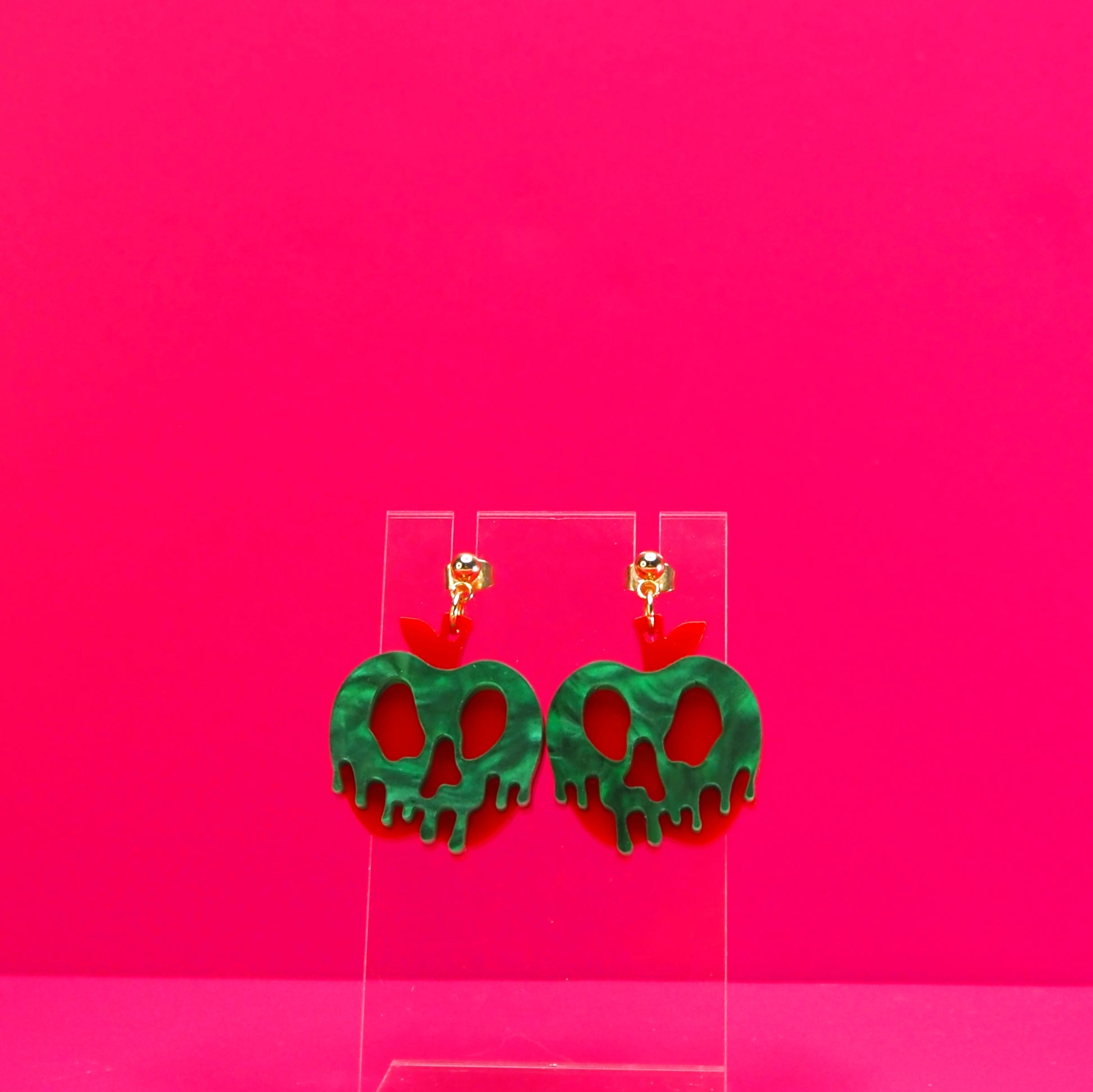 Red apple with green pearl goo earrings on a clear display against a pink background.