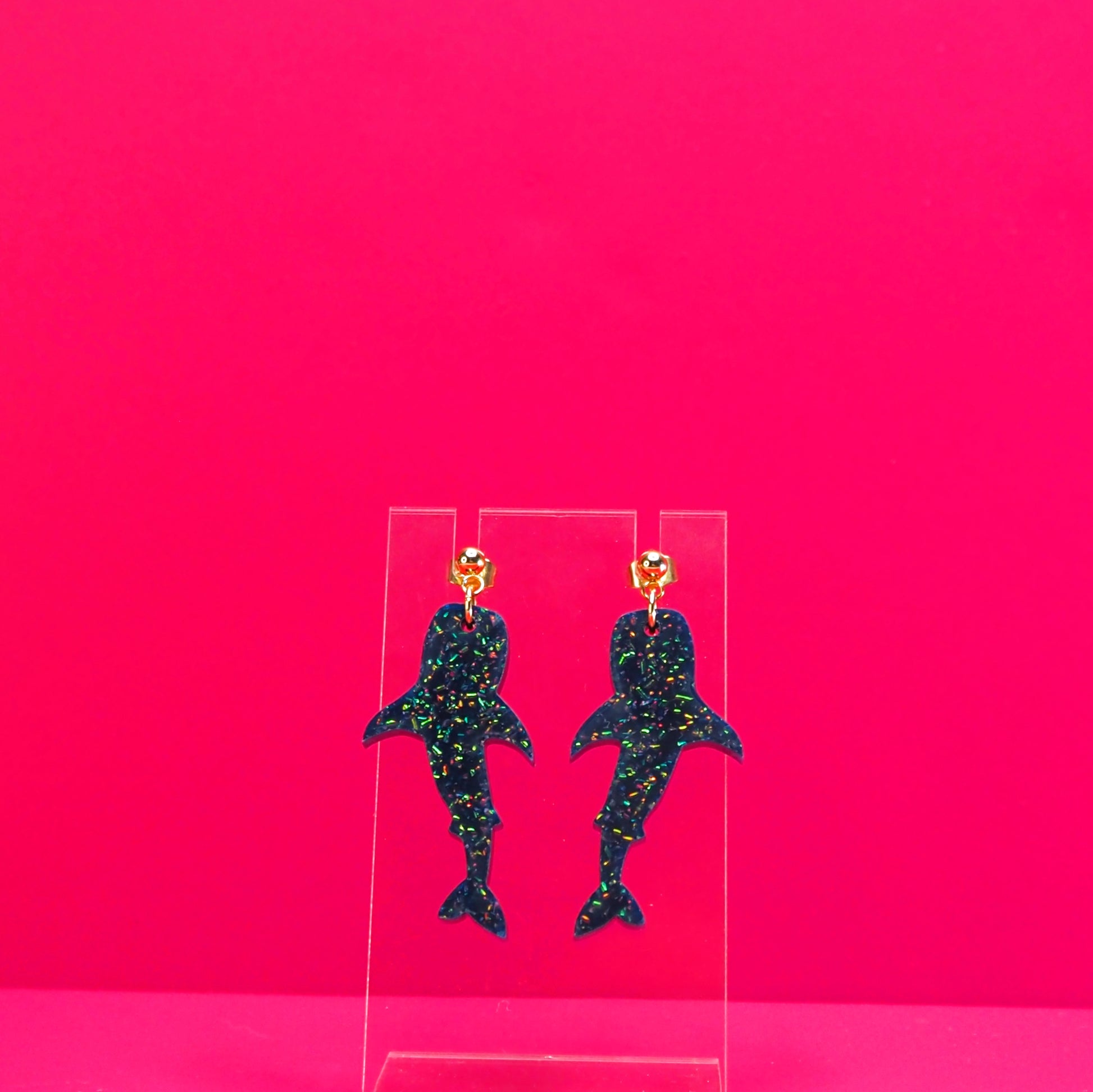 Navy blue glittery whale shark earrings on a clear display against a pink background.
