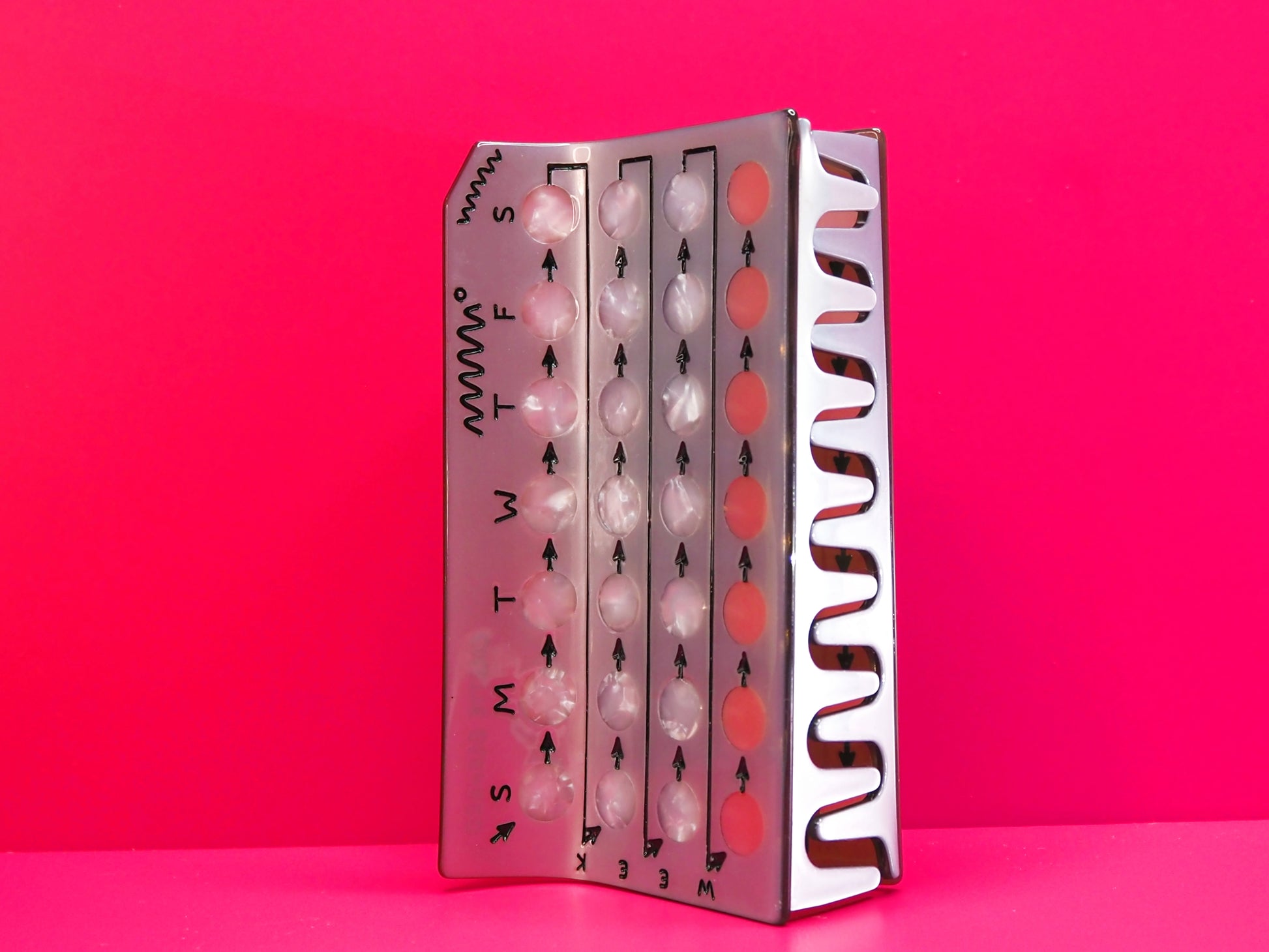 Silver birth control hair claw on its side against a pink background.
