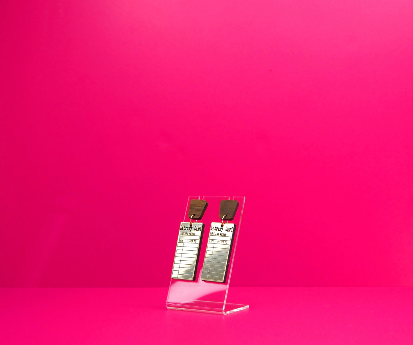 Gold rectangle library card earrings with wood accents on a clear display at an angle against a pink background.