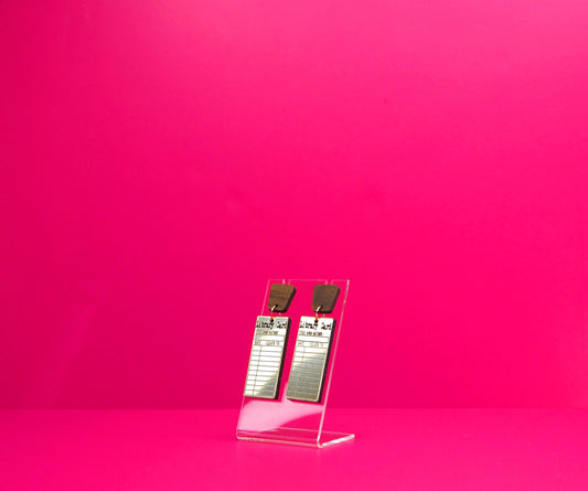Gold rectangle library card earrings with wood accents on a clear display at an angle against a pink background.