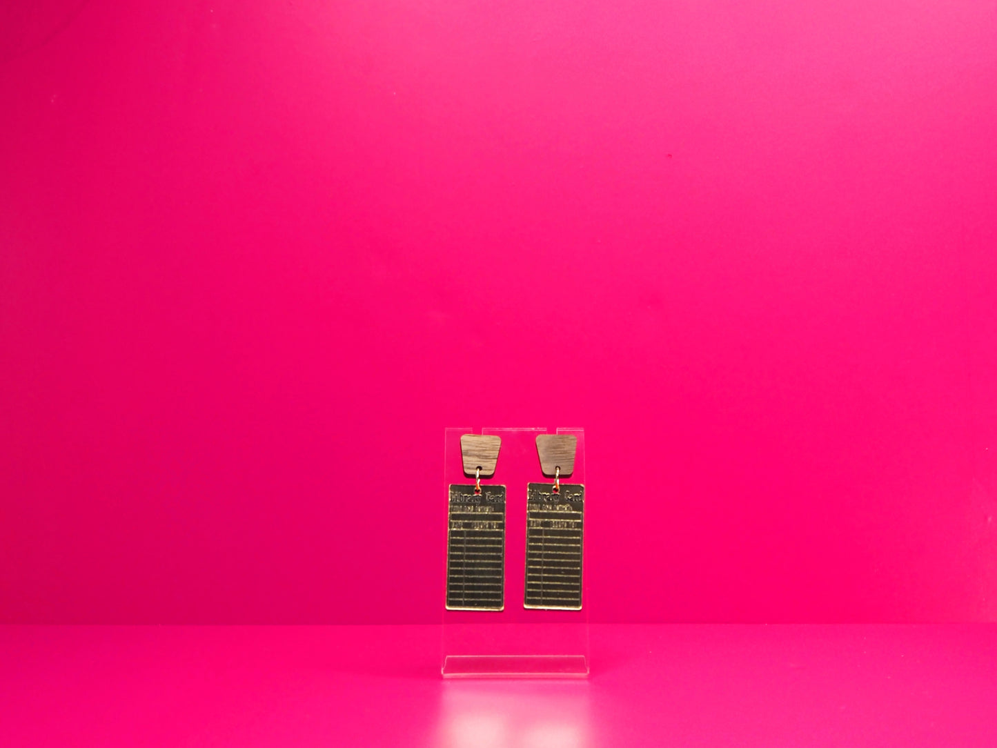 Gold rectangle library card earrings with wood accents on a clear display against a pink background.