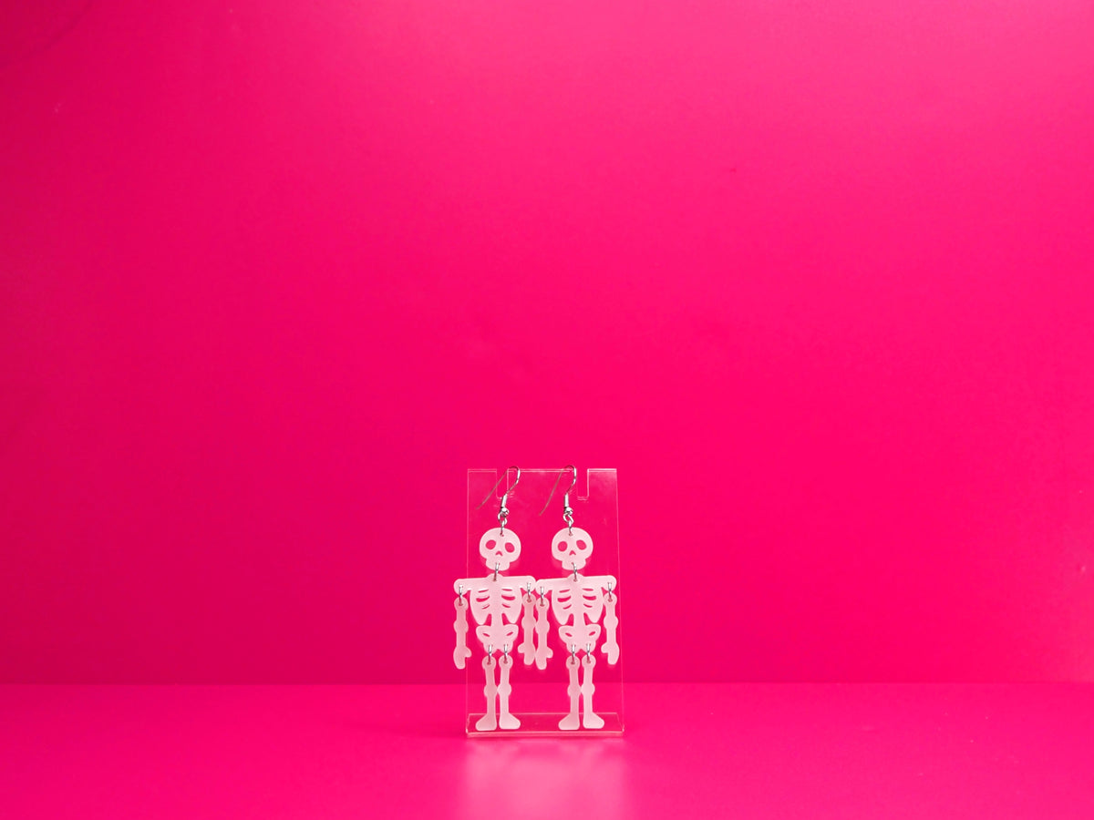 White acrylic skeleton earrings on a clear display against a pink background.