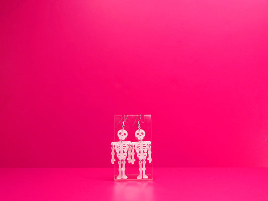White acrylic skeleton earrings on a clear display against a pink background.