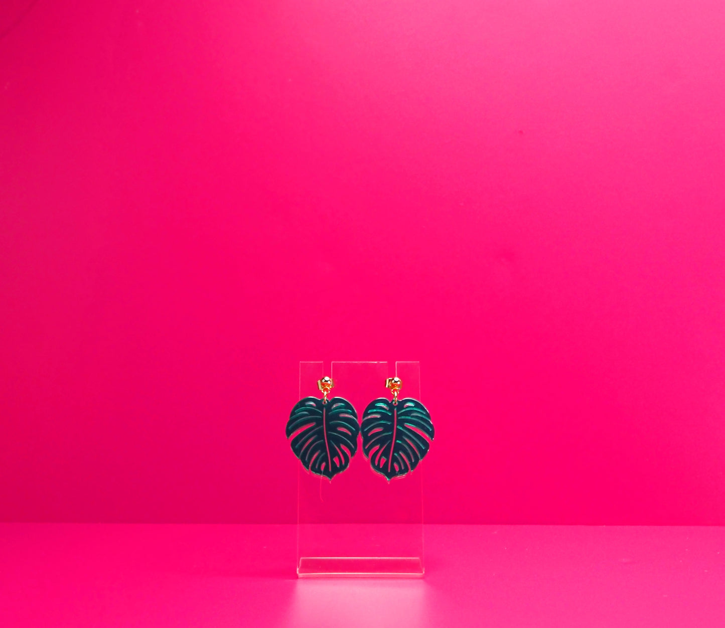 Green monstera leaf earrings on a clear display against a pink background.