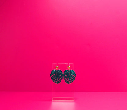 Green monstera leaf earrings on a clear display against a pink background.