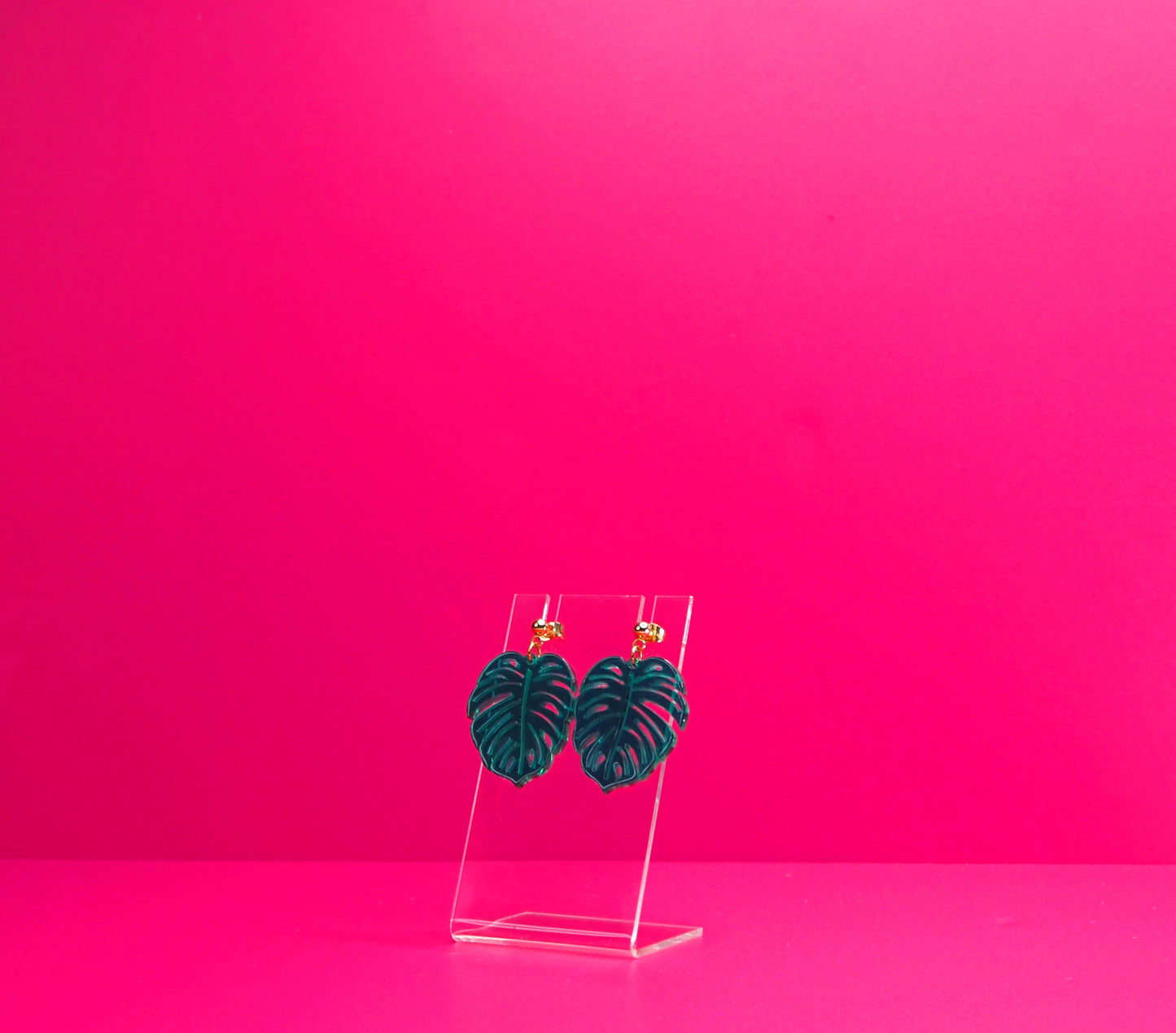 Green monstera leaf earrings on a clear display at an angle against a pink background.