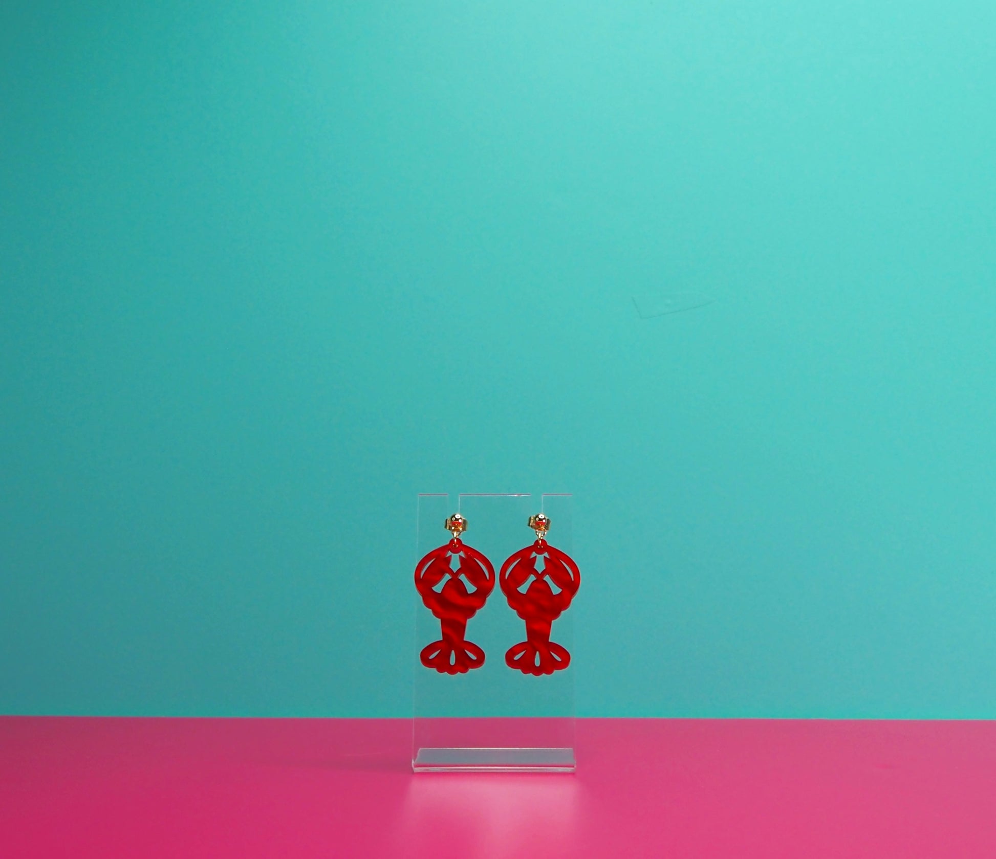 Red marbled lobster earrings on a clear display against a blue and pink background.