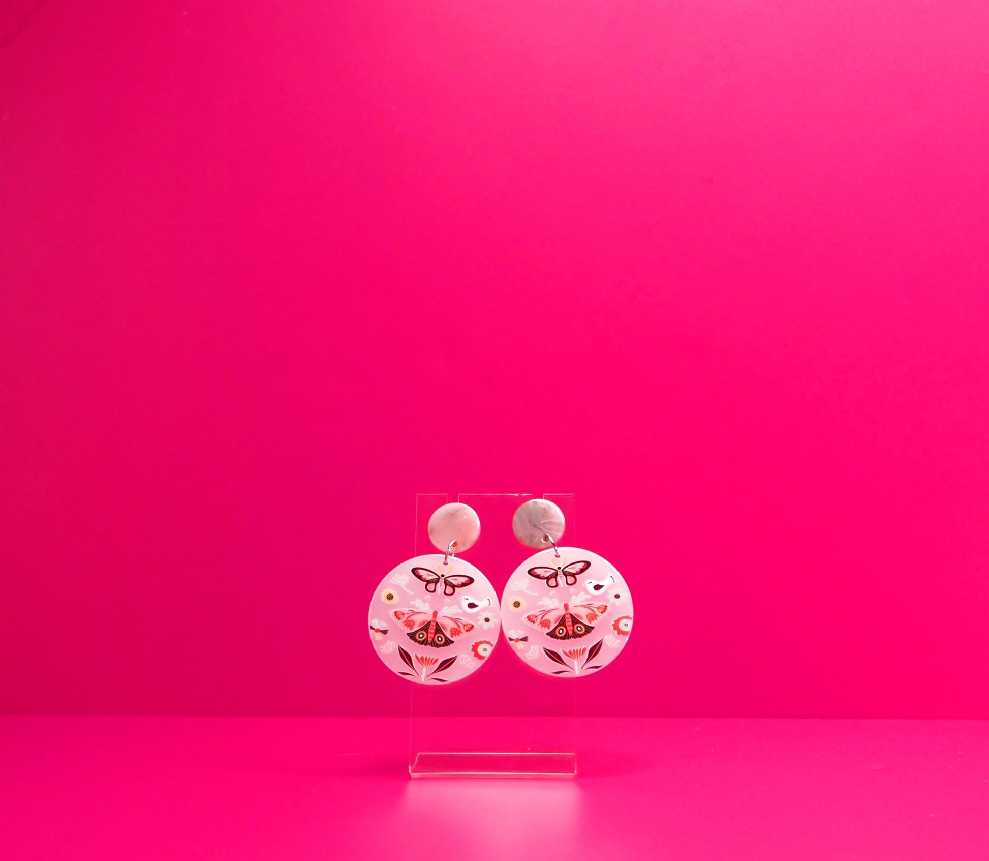 Round pink earrings with moths on a clear display against a pink background.