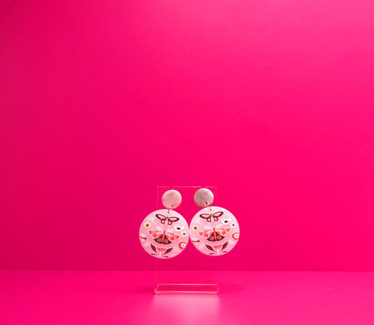 Round pink earrings with moths on a clear display against a pink background.