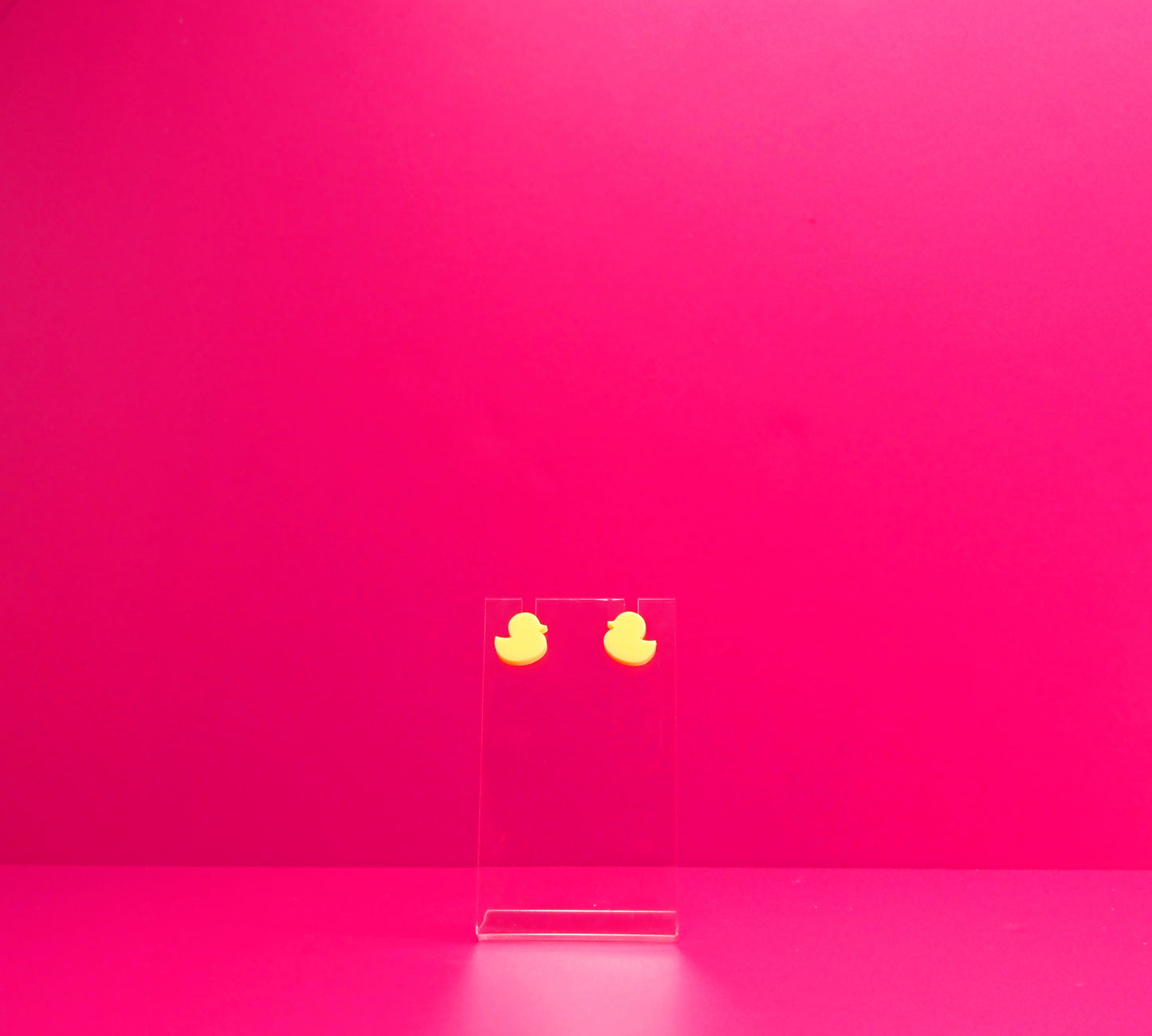 Yellow duck earrings on a clear display against a pink background.