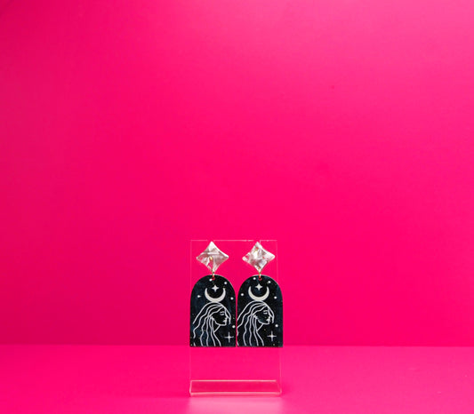 Celestial woman earrings with a crescent moon on a blue acrylic on a clear display against a pink background.
