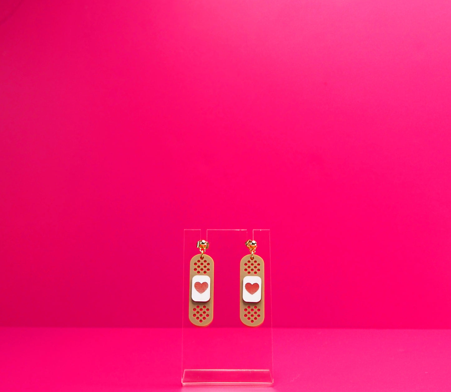 Brown bandage earrings on a clear display against a pink background.