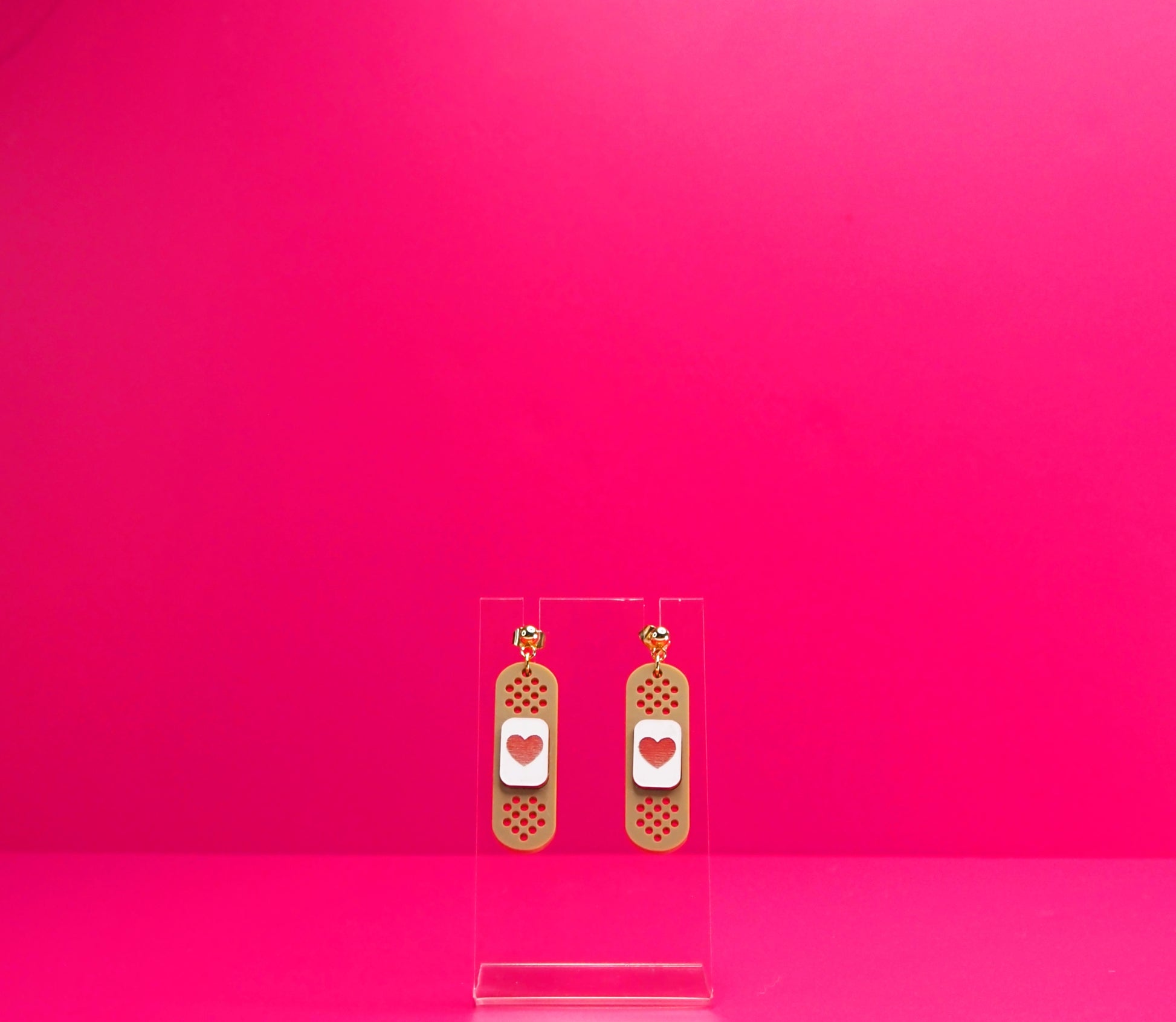 Brown bandage earrings on a clear display against a pink background.