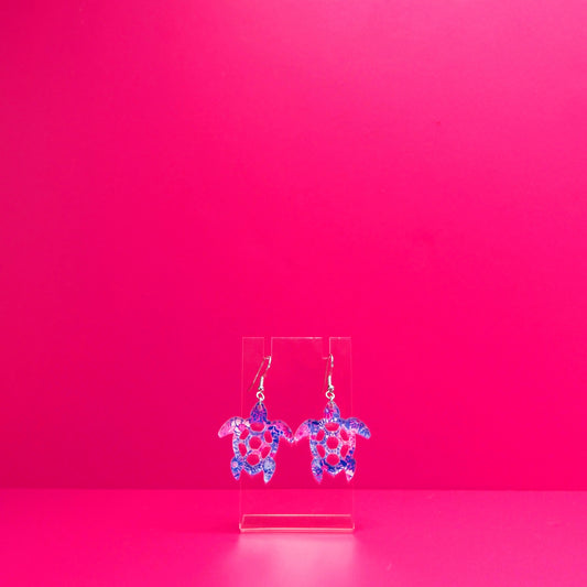 Purple  blue, pink, white floral sea turtle earrings on a clear display against a pink background.