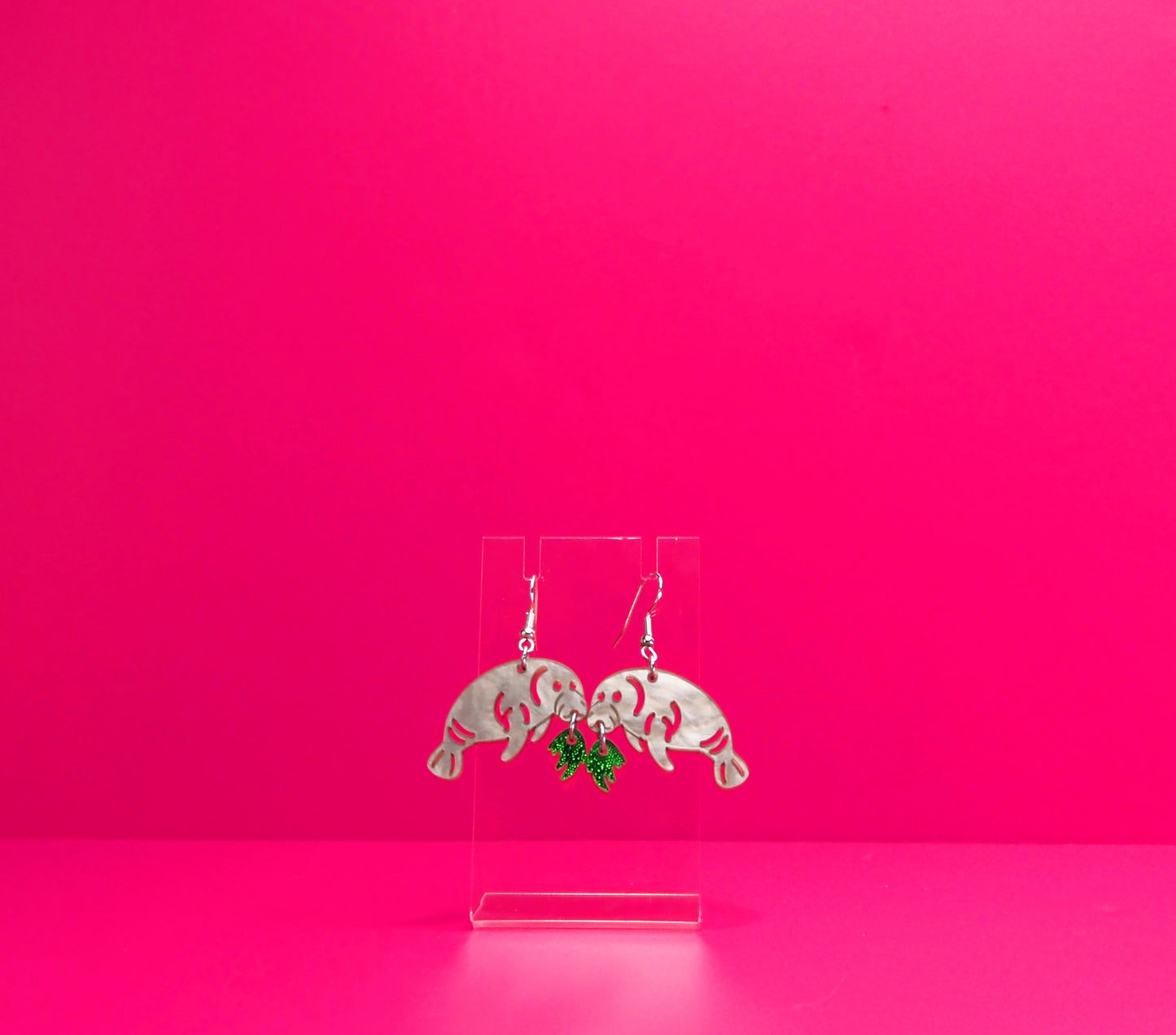 Grey manatee earrings eating green kelp on a clear display against a pink background.