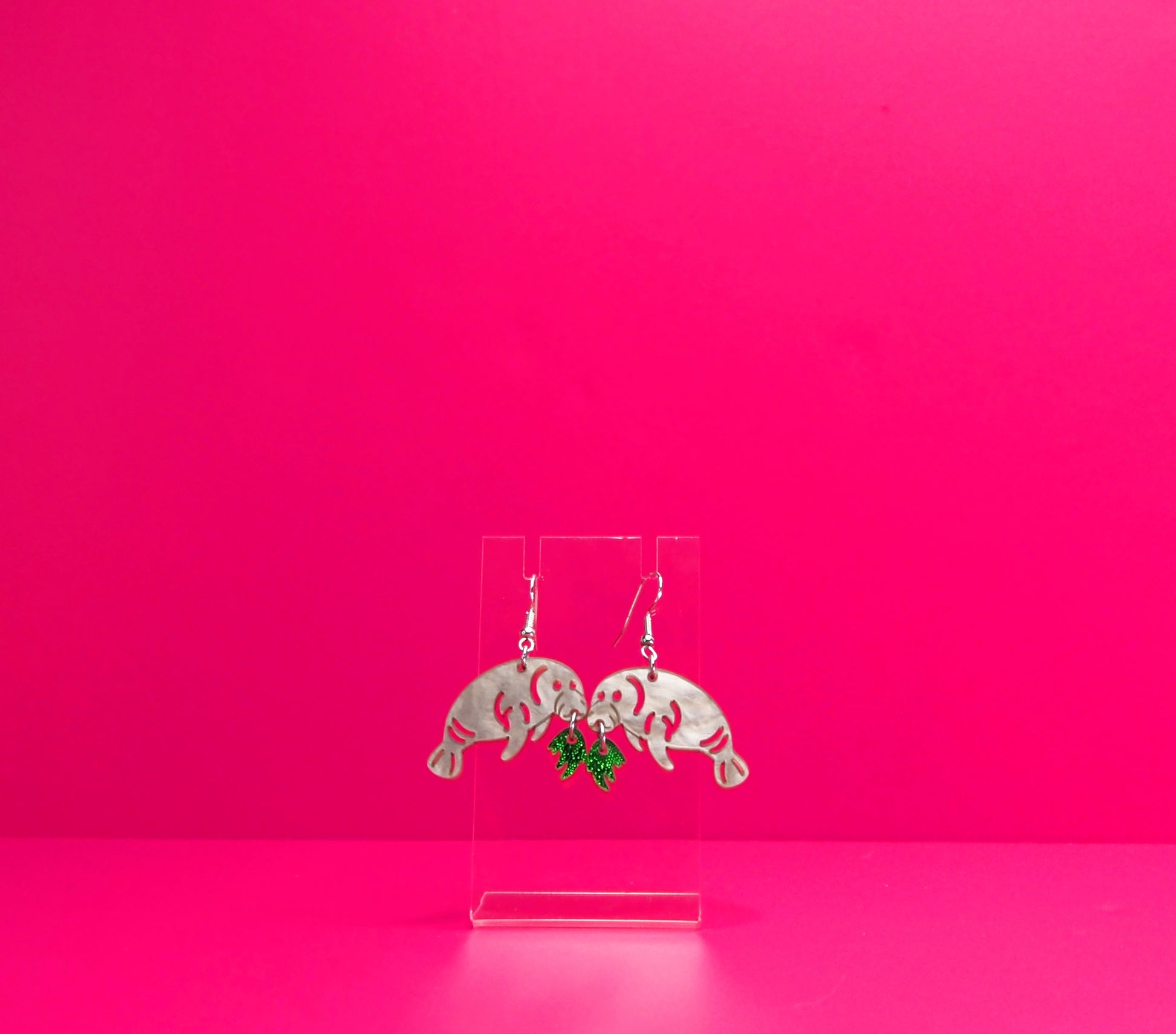 Grey manatee earrings eating green kelp on a clear display against a pink background.