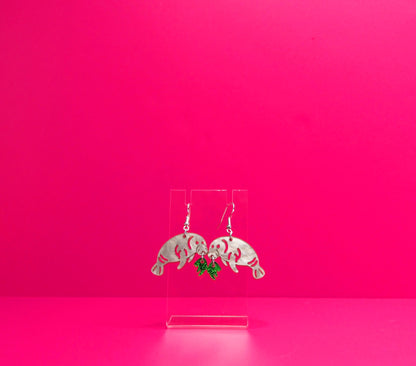 Grey manatee earrings eating green kelp on a clear display against a pink background.