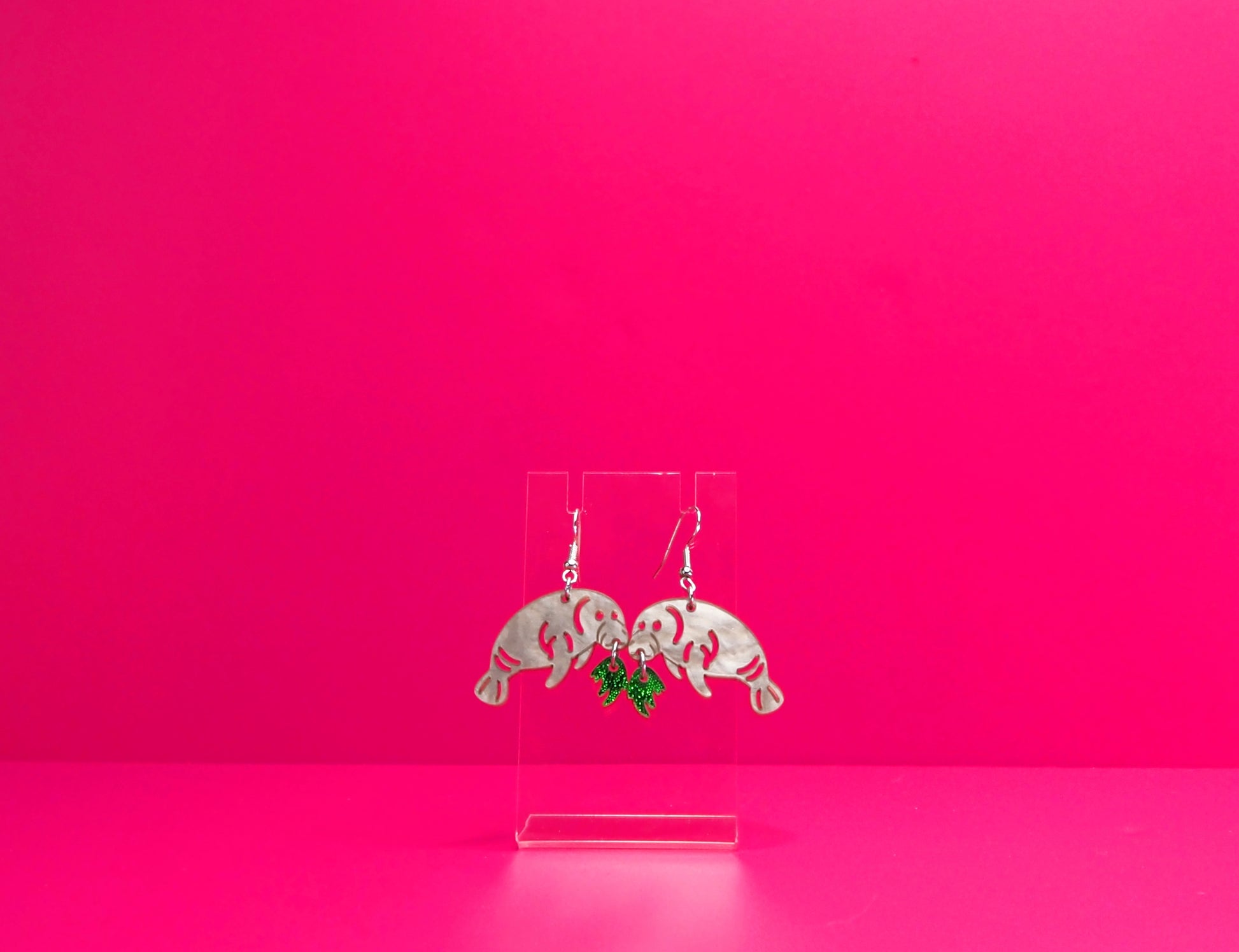 Grey manatee earrings eating green kelp on a clear display against a pink background.