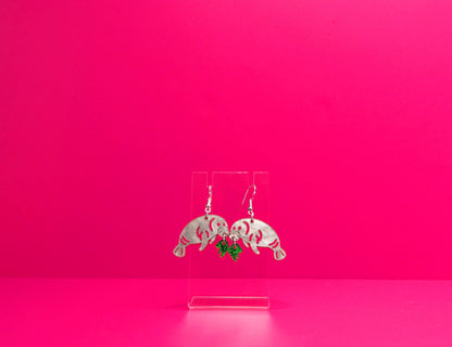 Grey manatee earrings eating green kelp on a clear display against a pink background.