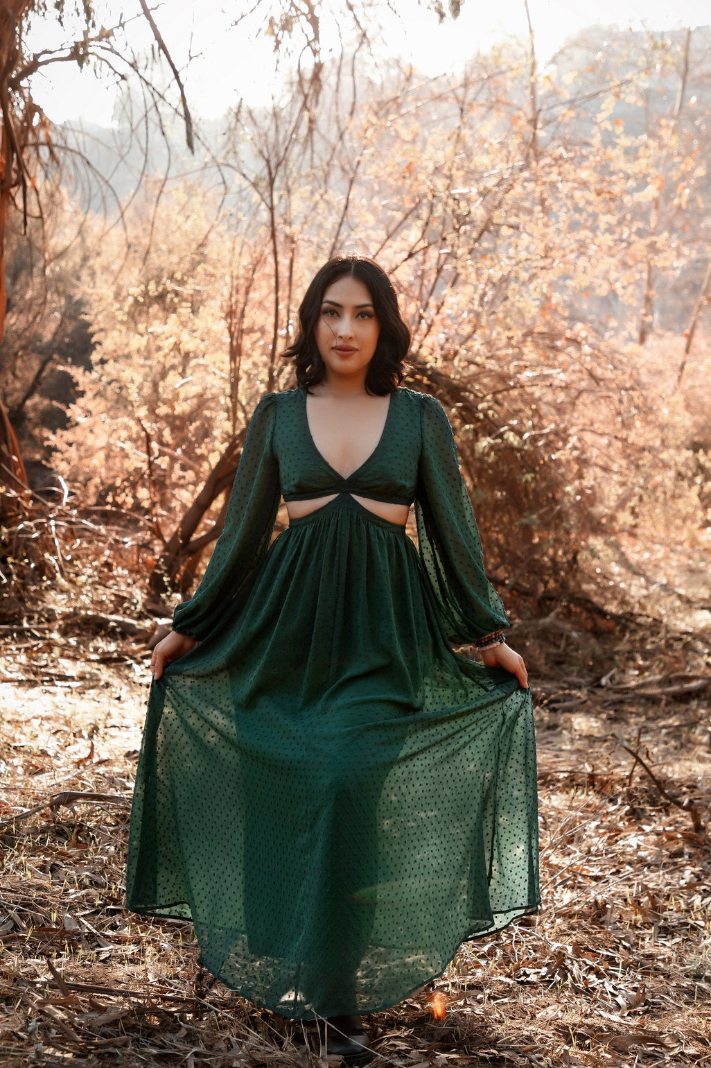 Female presenting model in the green  Feralune dress standing in a forest with dry leaves and branches.