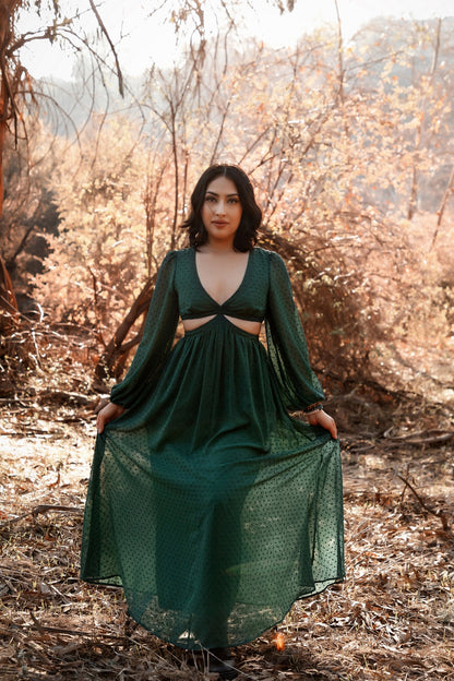 Female presenting model in the green  Feralune dress standing in a forest with dry leaves and branches.