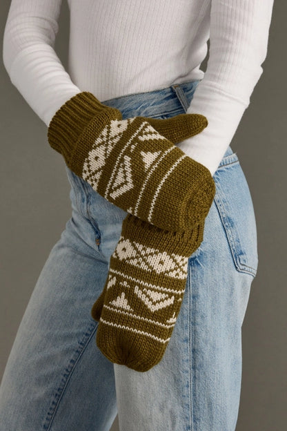 Green knitted mittens with a geometric pattern worn by a person in a white long-sleeve shirt and blue jeans.